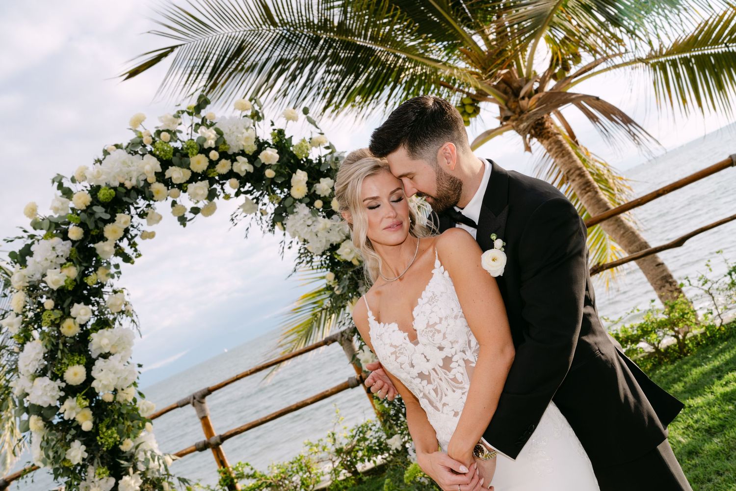 Bride and groom embrace tenderly under a lush white floral arch with palm trees and ocean backdrop.