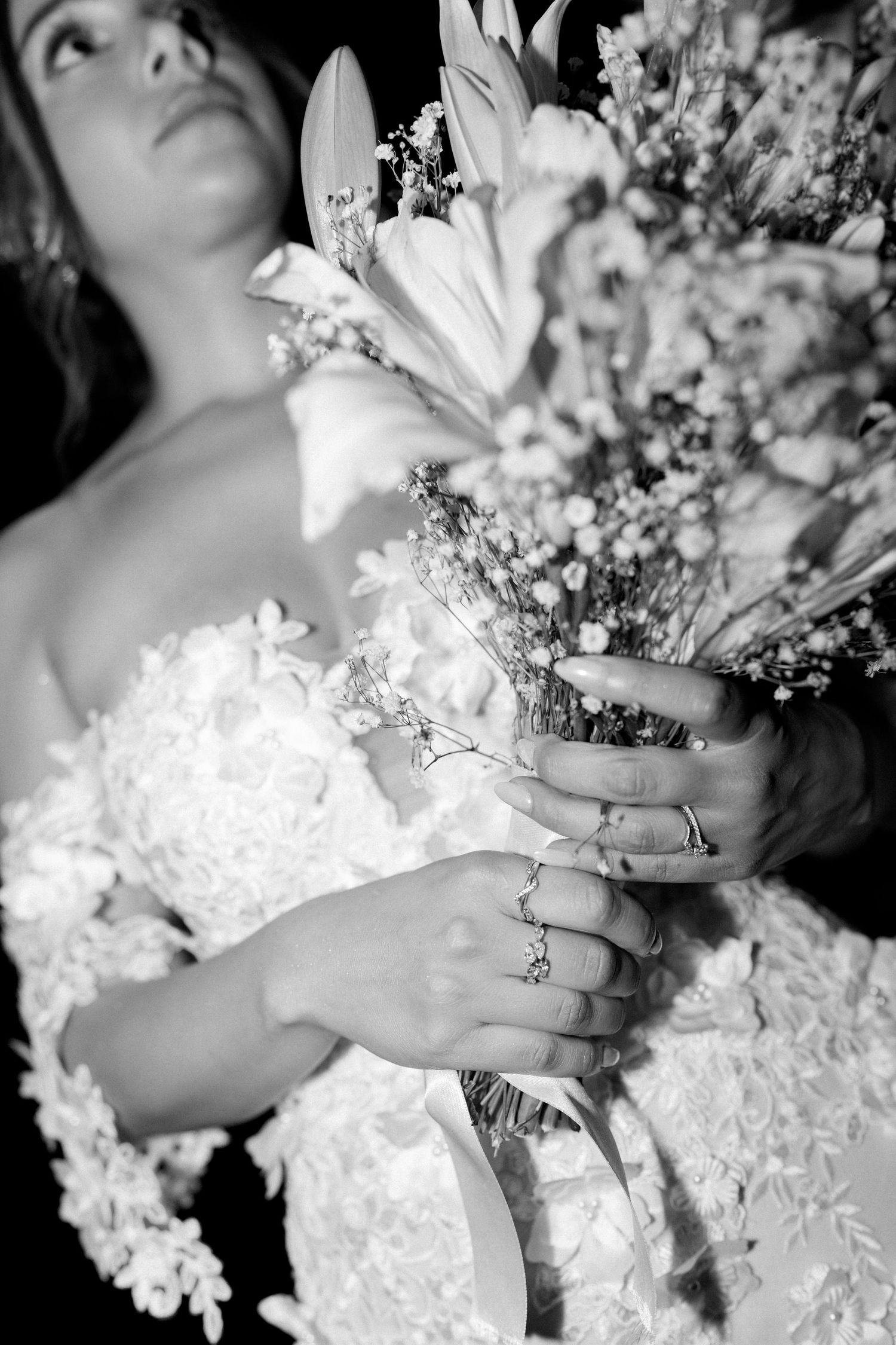 Black and white close-up of bride holding floral bouquet wearing lace gown and diamond rings.