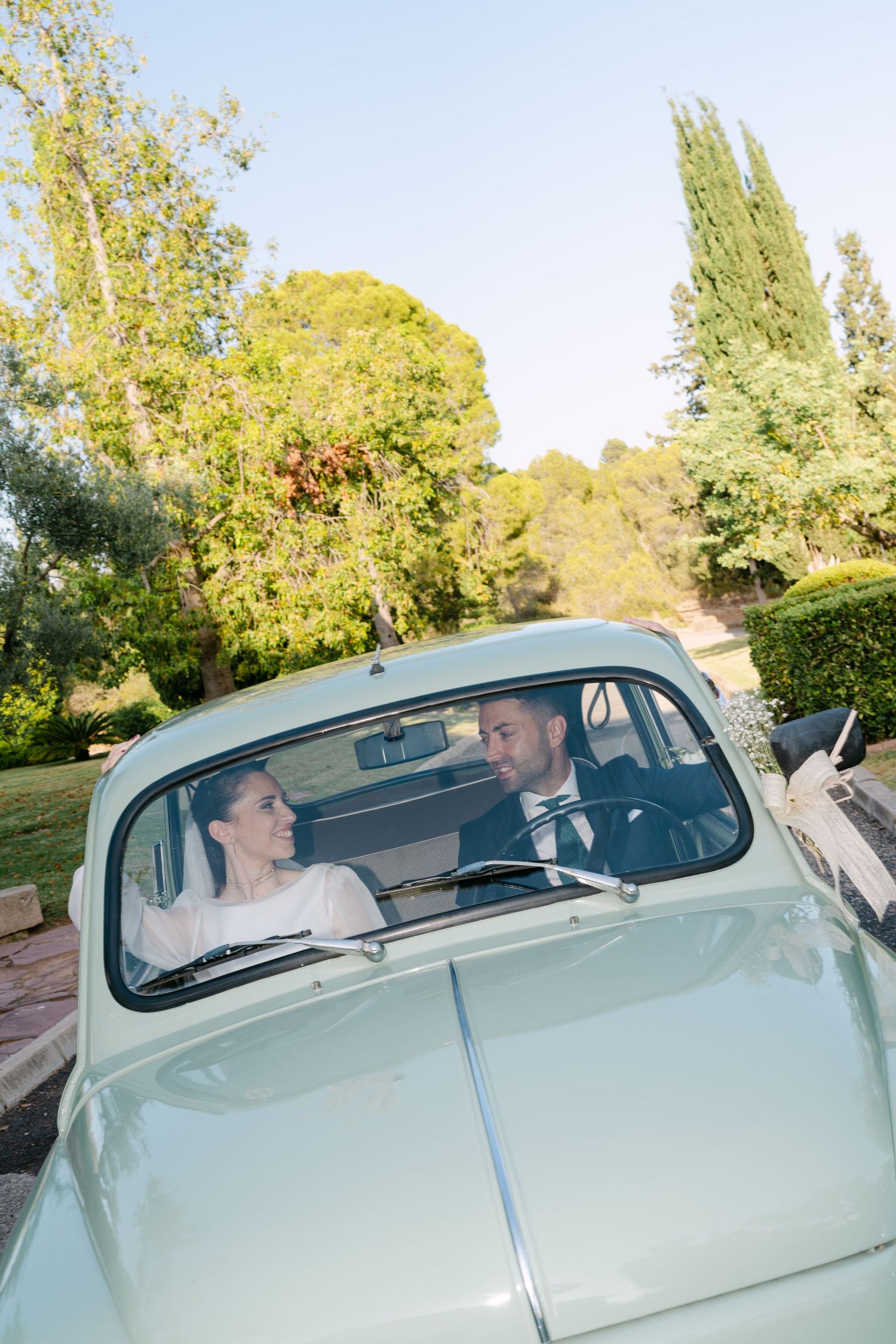 Newlyweds smile at each other inside a vintage mint green car surrounded by lush greenery.