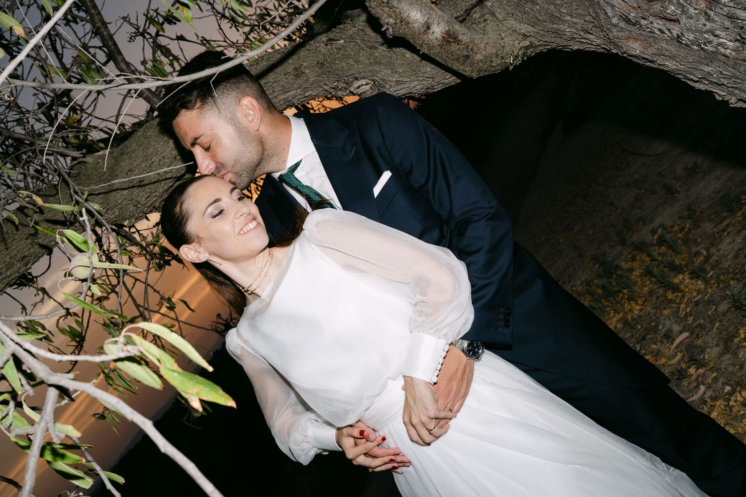 Romantic bride and groom cuddle closely together beneath rustic tree branches at night.