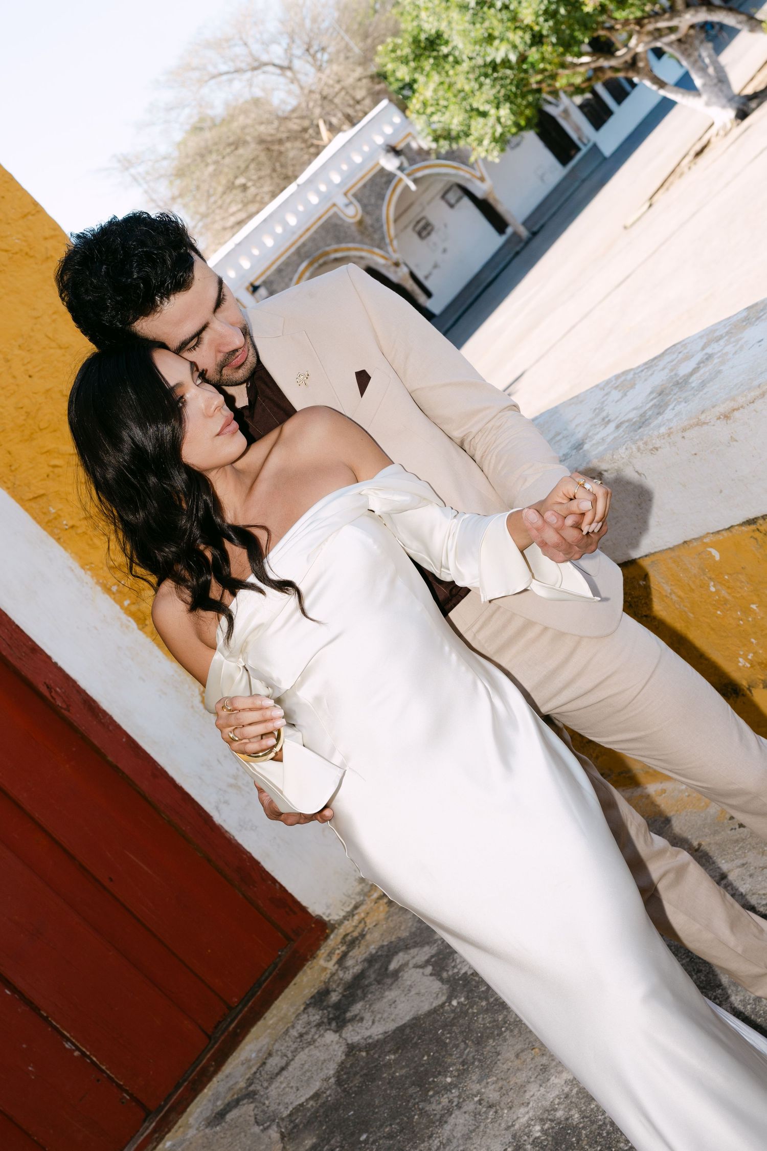 Groom in beige suit embraces bride in white off-shoulder gown against yellow wall near red door.