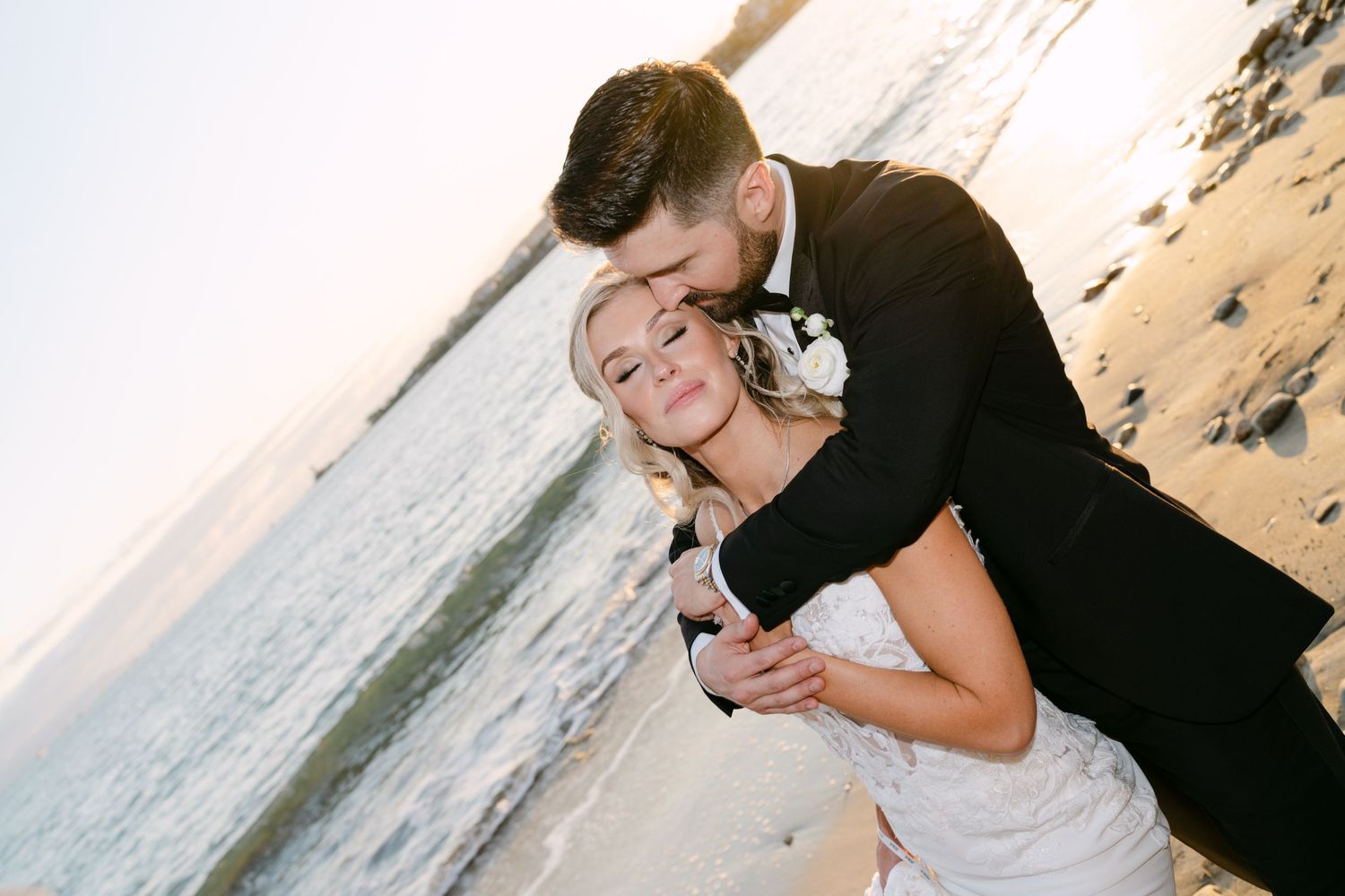 Groom tenderly kisses bride on the cheek on a sunlit beach, bride smiling in strapless wedding dress.