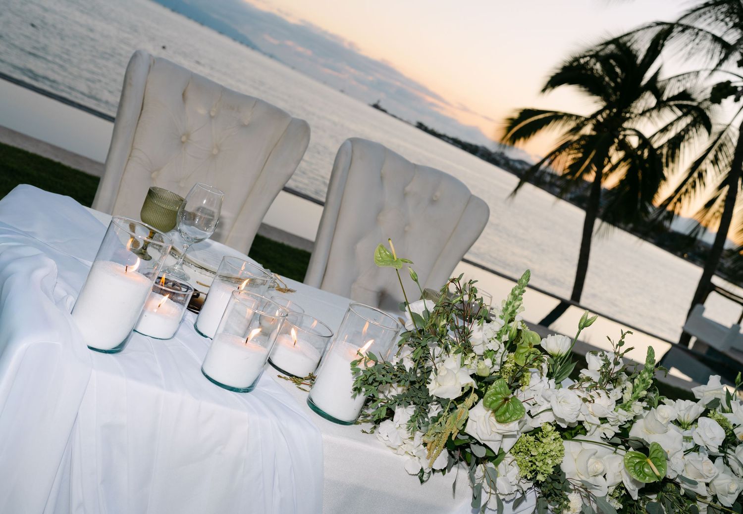 Elegant sweetheart table with white florals and candles overlooking ocean at sunset.