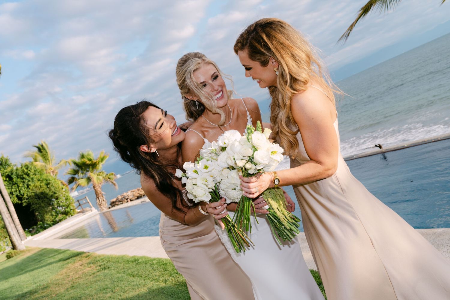 Bride and two bridesmaids laughing together with white floral bouquet at beachside wedding.