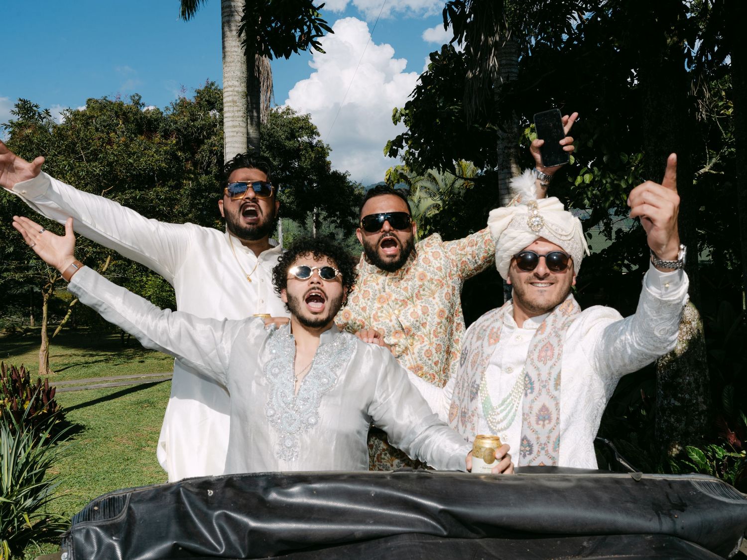 Groom in white sherwani and turban celebrating with groomsmen in matching white outfits outdoors.