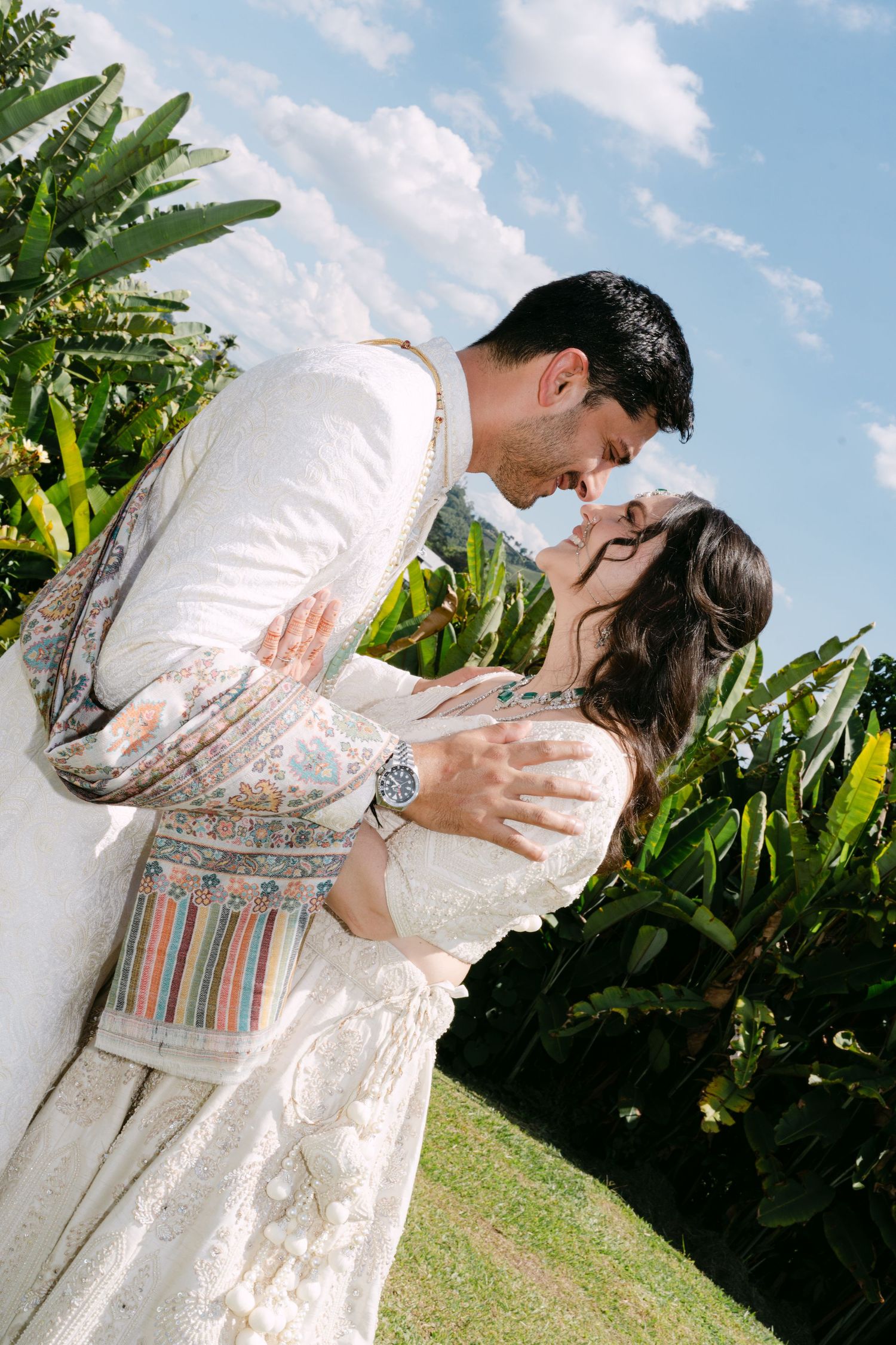 Couple in traditional Indian wedding attire share a romantic embrace surrounded by tropical greenery.