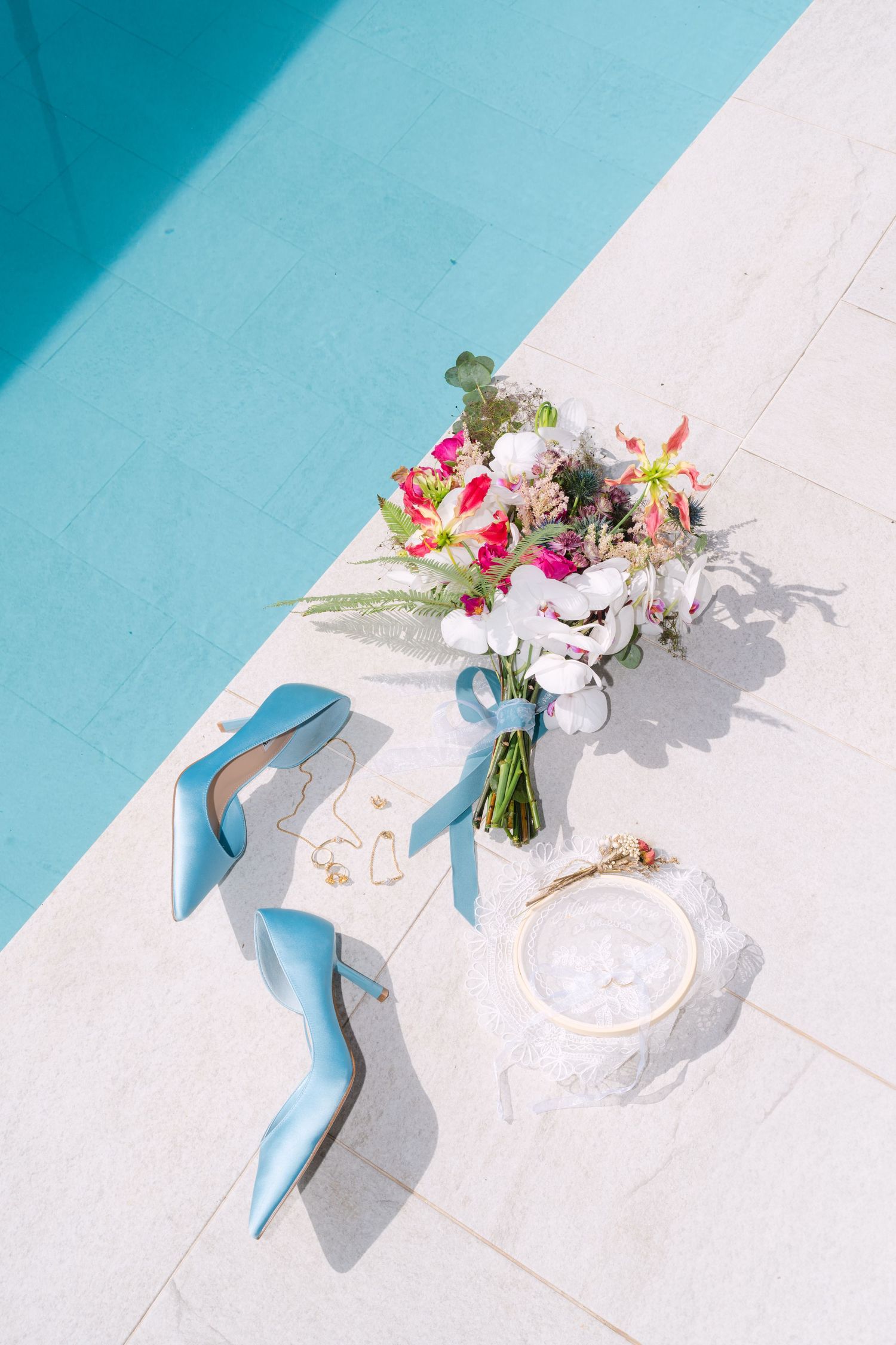 Blue bridal heels, floral bouquet, and gold jewelry accessories arranged by a poolside.