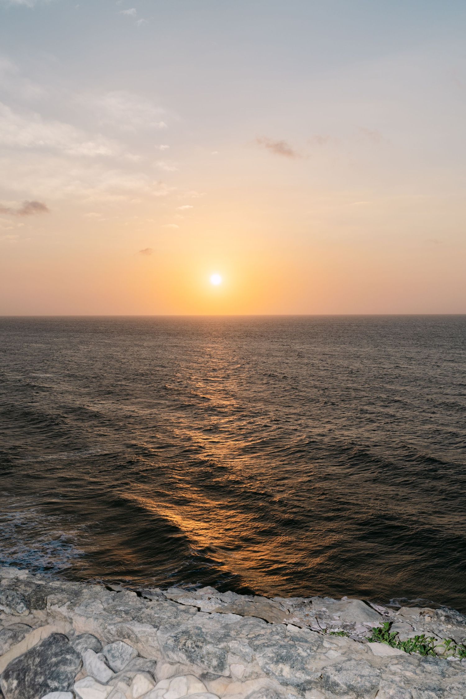 Stunning ocean sunset viewed from rocky cliffside with golden light reflecting on dark water.
