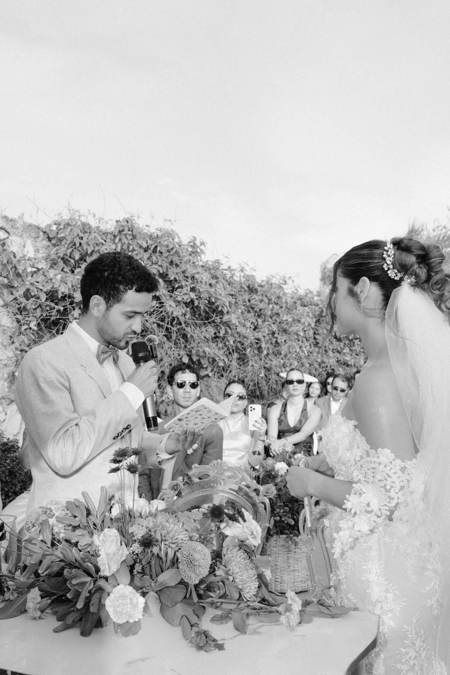 Black and white photo of groom reading vows at outdoor wedding ceremony with floral arrangements.
