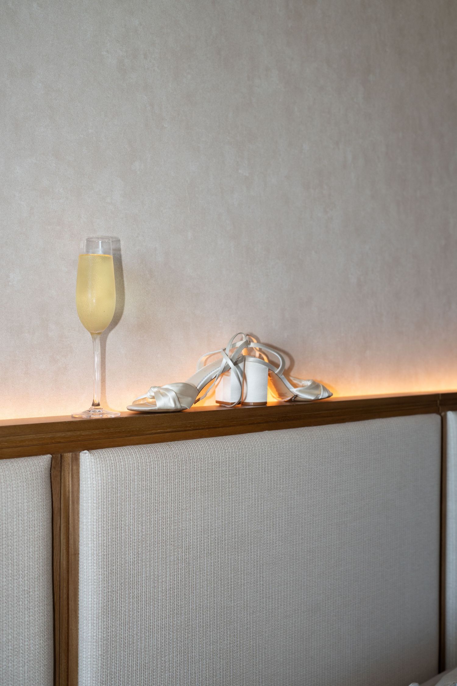 White bridal heels and a champagne flute displayed on an illuminated upholstered headboard.