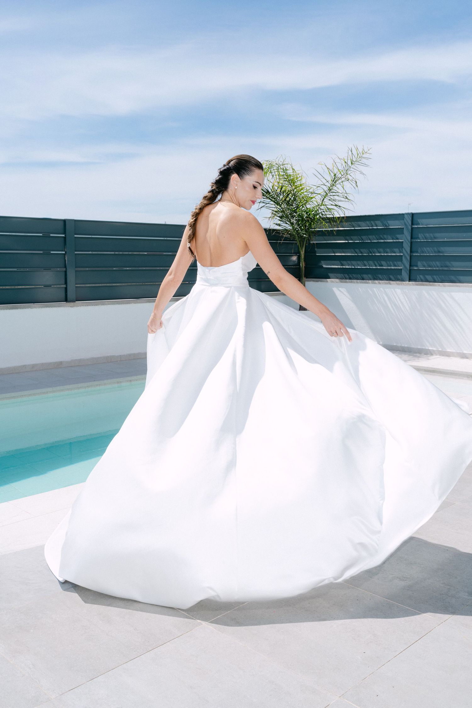 Bride in a flowing white strapless ball gown twirling near a pool under a blue sky.