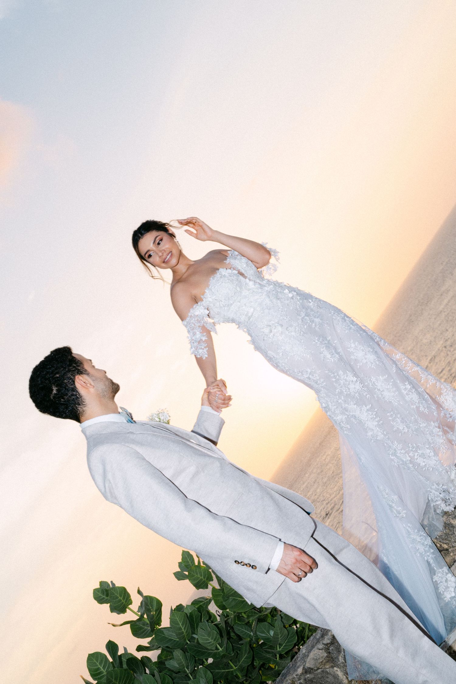 Bride and groom hold hands at sunset beach wedding, she in lace gown, he in light gray suit.