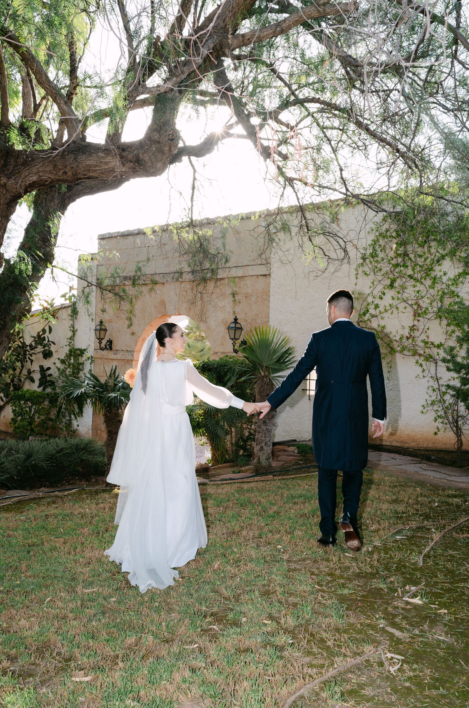 Bride and groom holding hands walking toward elegant venue under large tree on their wedding day.