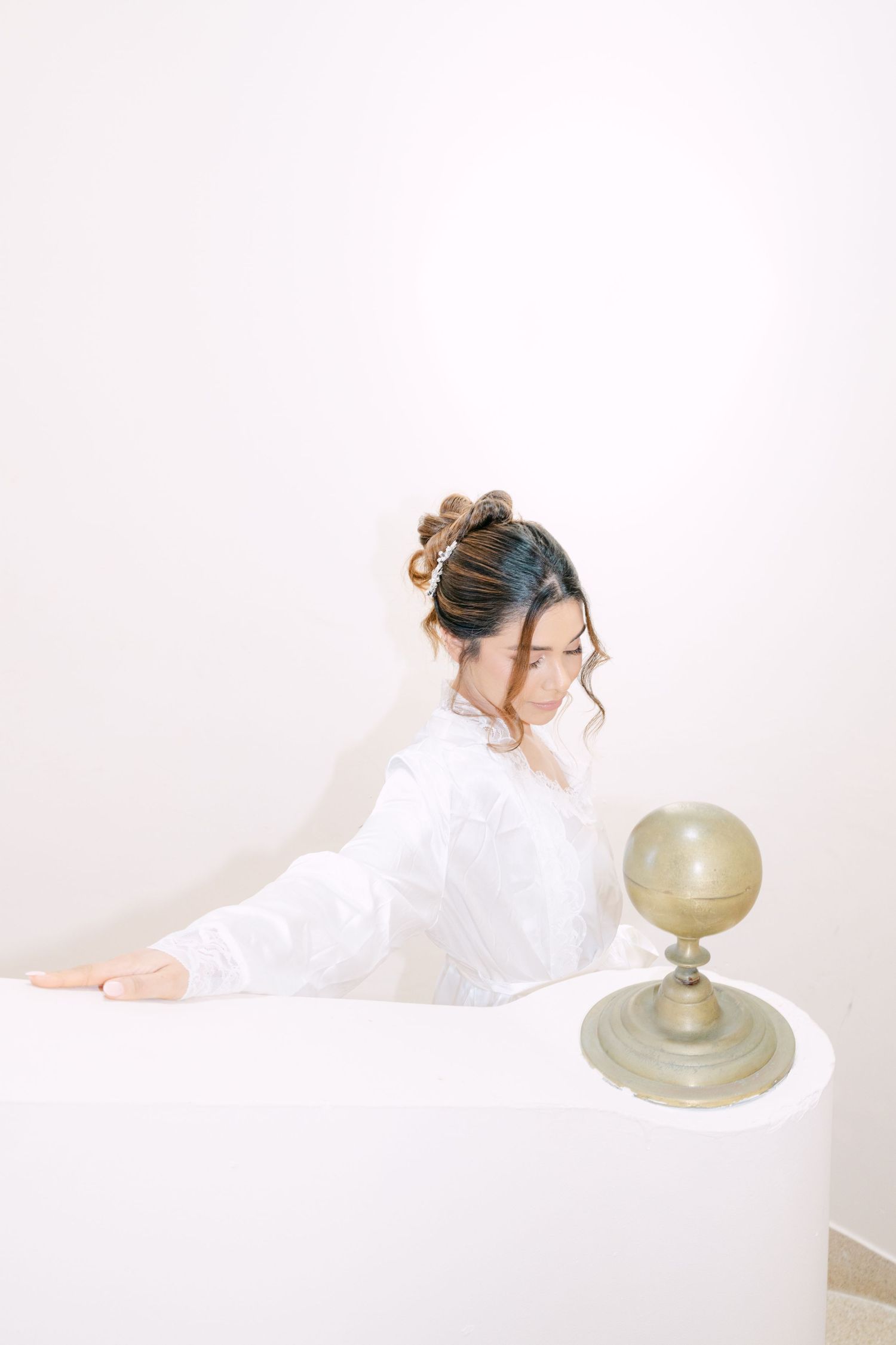 Beautiful bride in white robe with elegant updo hairstyle posing near decorative globe on white staircase.