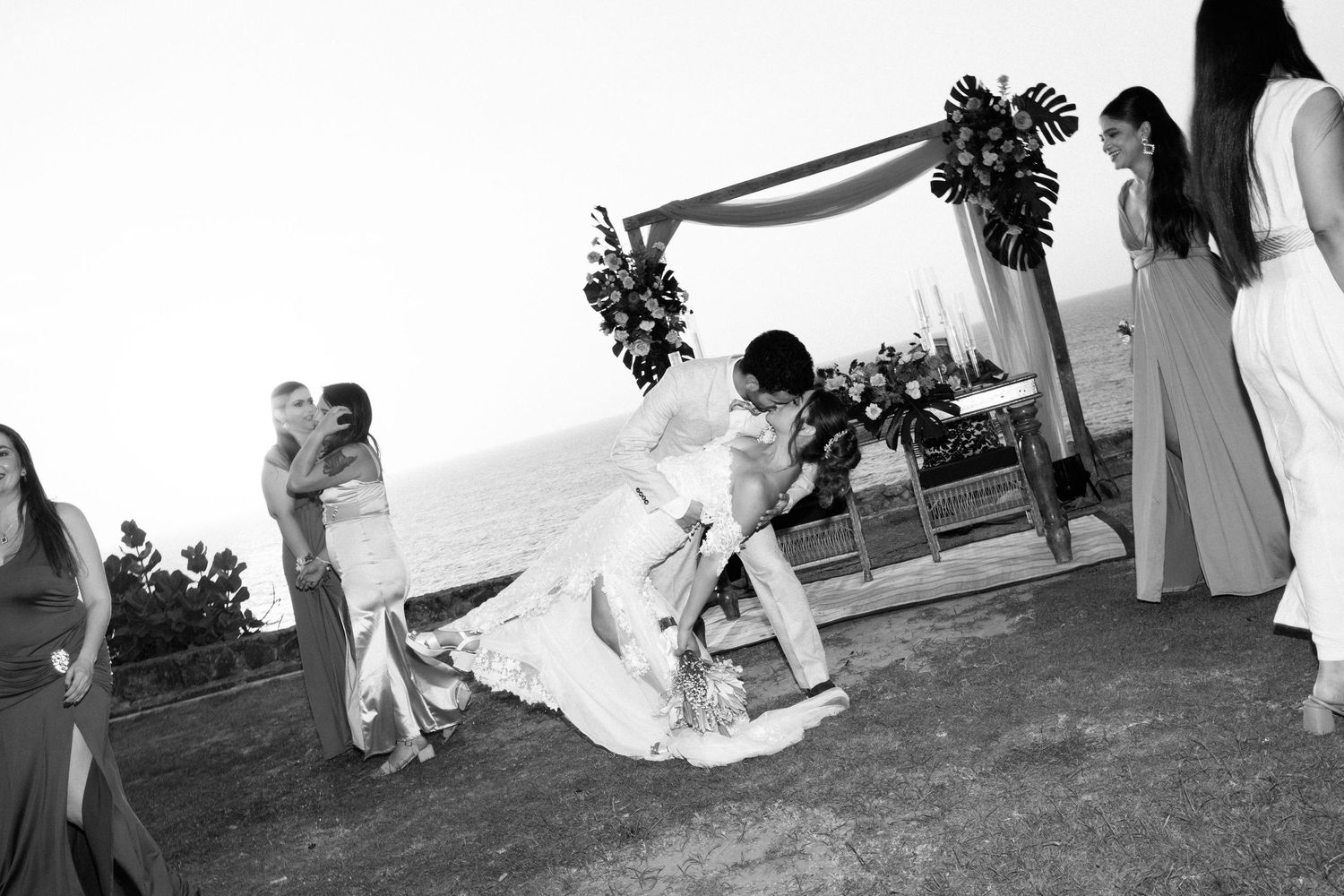 Black and white photo of bride adjusting dress near floral arch with ocean view backdrop.