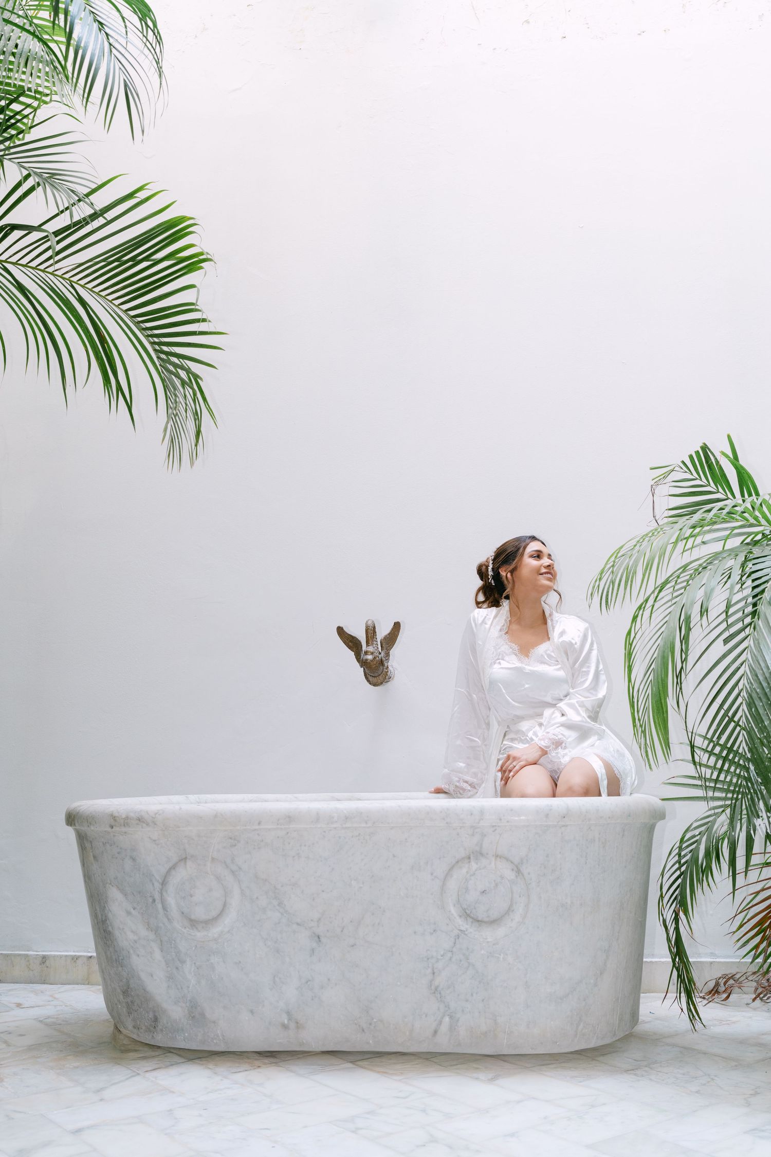 Elegant bride in white robe sitting on marble bathtub surrounded by lush tropical palm plants.