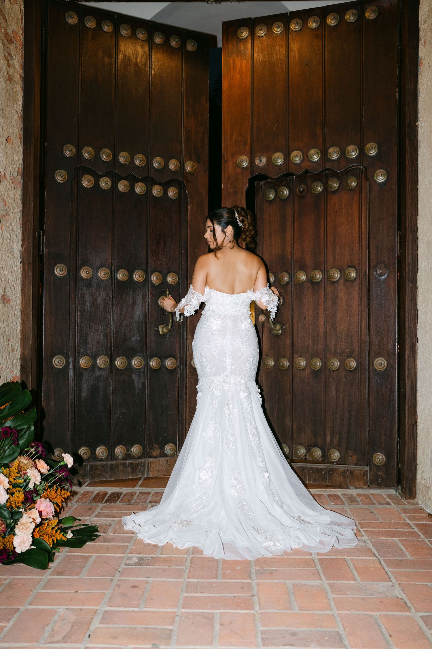 Bride in off-shoulder lace mermaid gown stands before ornate studded wooden doors with floral arrangement nearby.