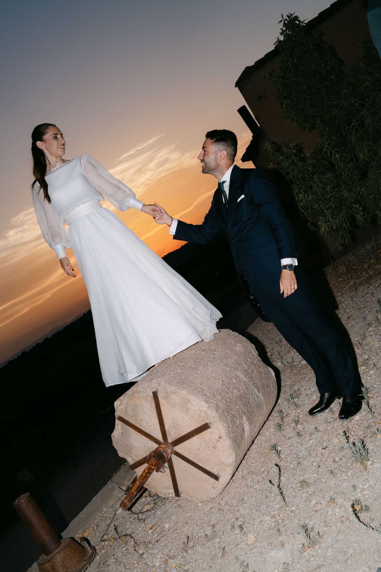 Bride stands on stone pedestal holding grooms hand at sunset during outdoor wedding photos.