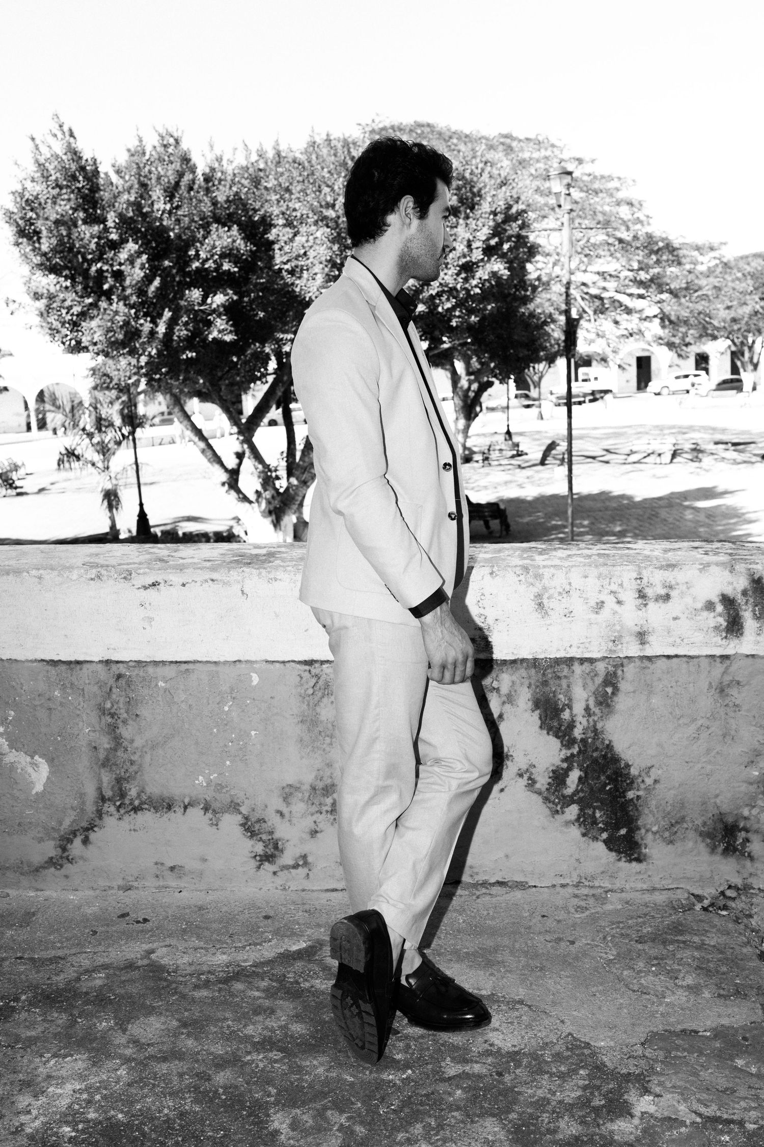 Dapper groom in light beige suit stands in profile against stone wall with trees in black and white photo.