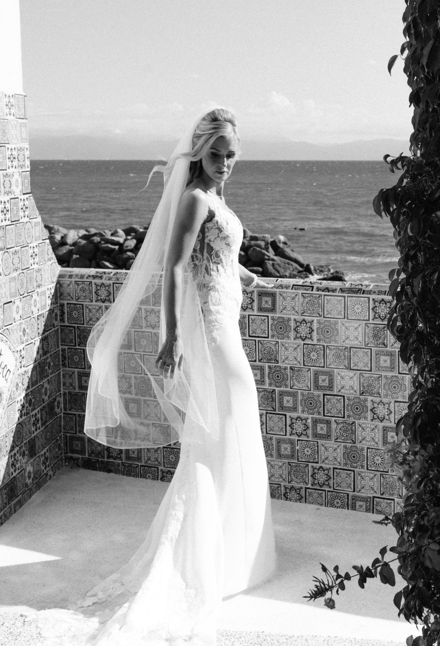 Black and white photo of a blonde bride in a fitted gown with veil by a tiled ocean wall.