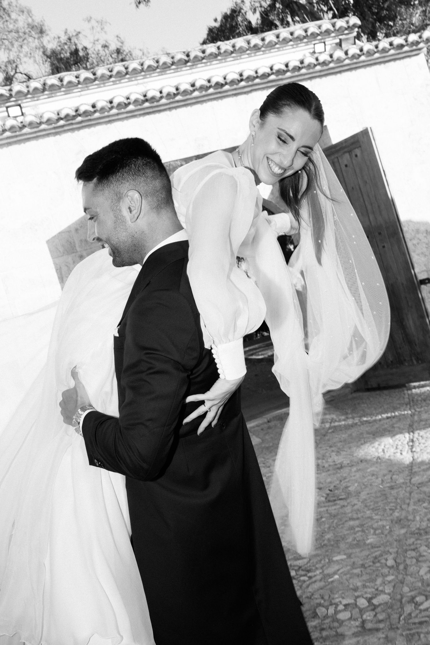 Black and white photo of groom carrying laughing bride on his back outside a white Spanish villa.