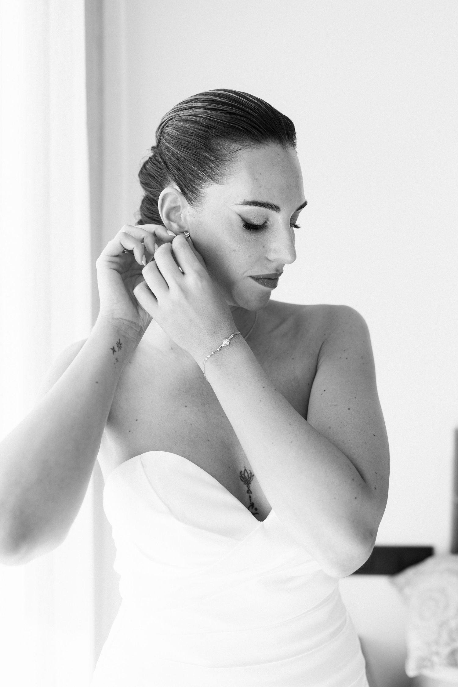 Black and white photo of a bride putting on earrings in a strapless wedding dress.