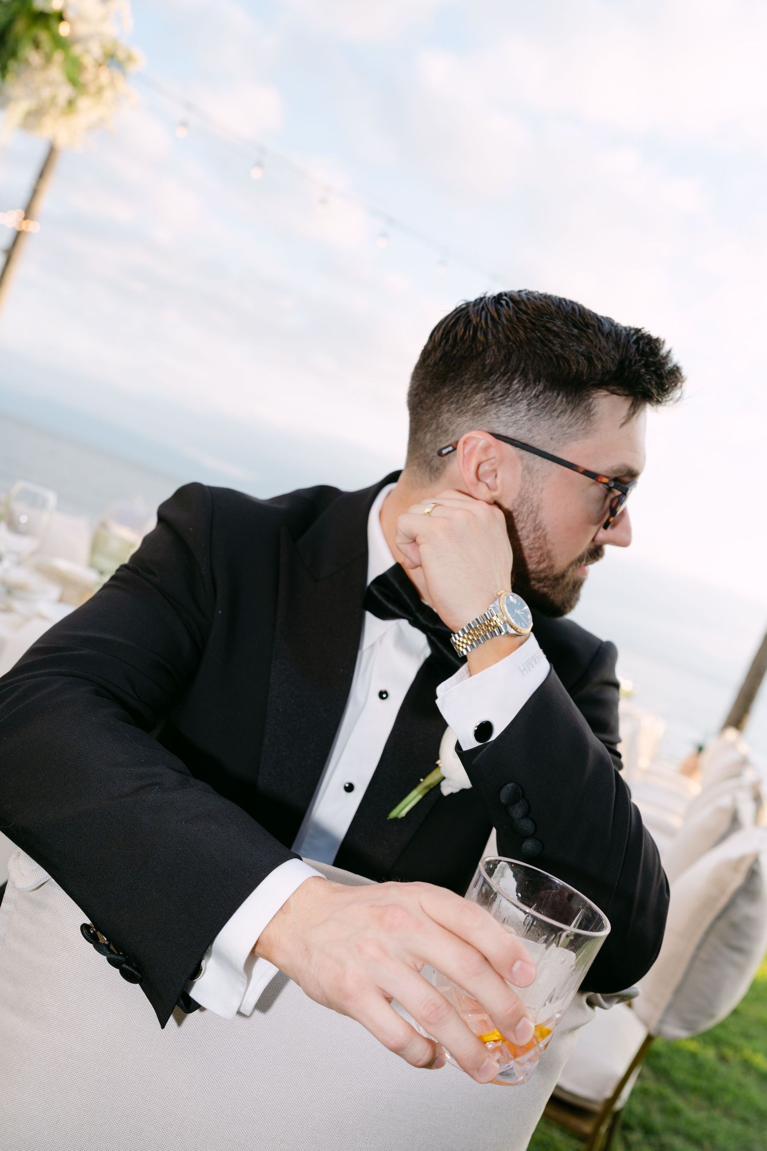 Dapper groom in black tuxedo holding whiskey glass at elegant oceanfront wedding reception.