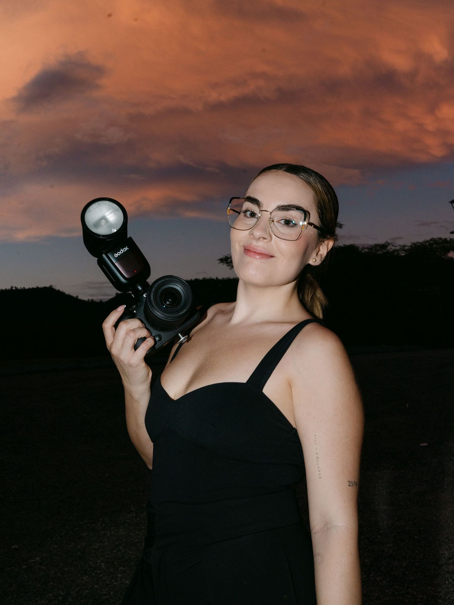 Female wedding photographer holds camera with flash at sunset against dramatic orange cloudy sky.