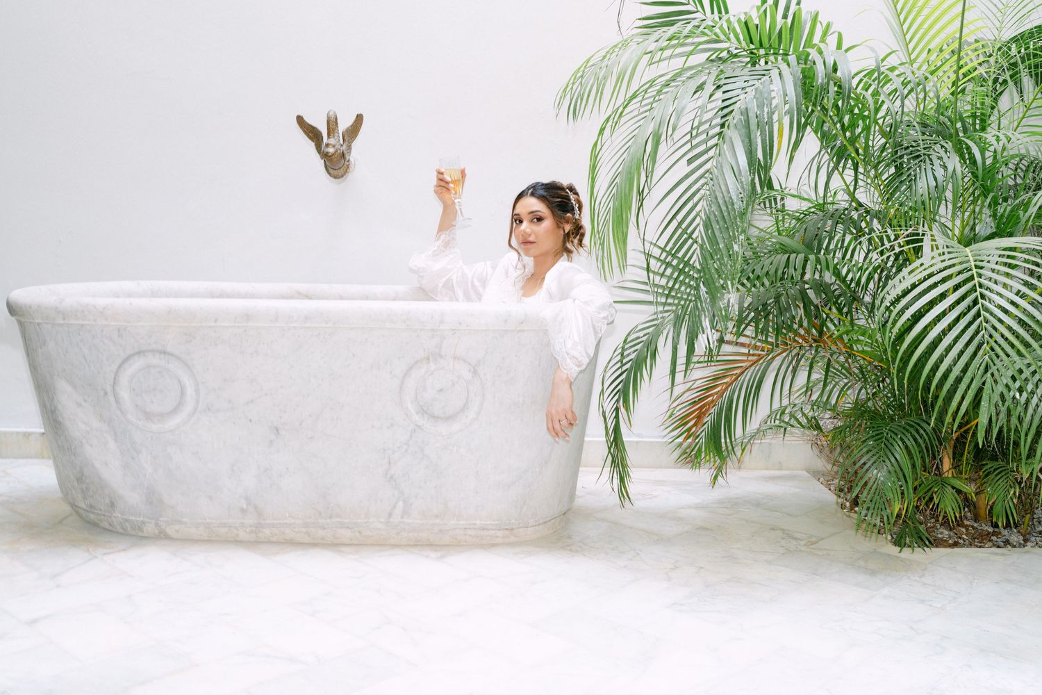 Joyful bride in white robe raising champagne glass in marble bathtub with tropical plants.
