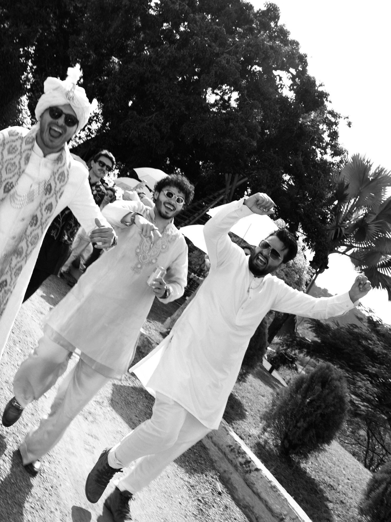 Black and white photo of groom and groomsmen in white kurtas joyfully walking outdoors at wedding celebration.