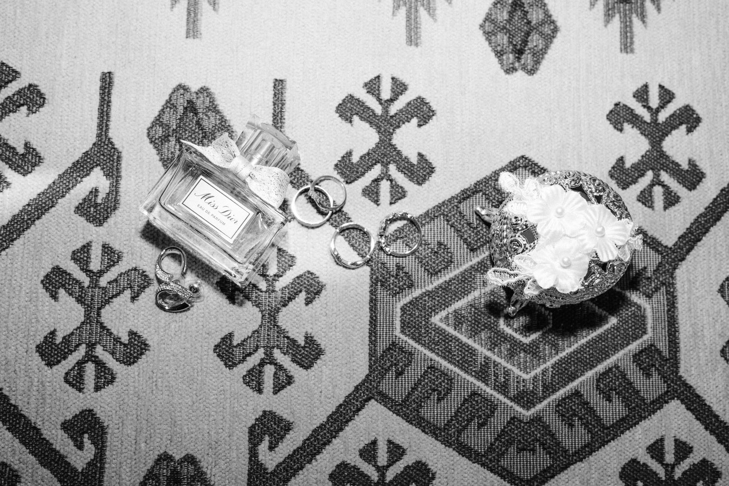Black and white flat lay of wedding perfume, rings, and floral accessories on patterned fabric.