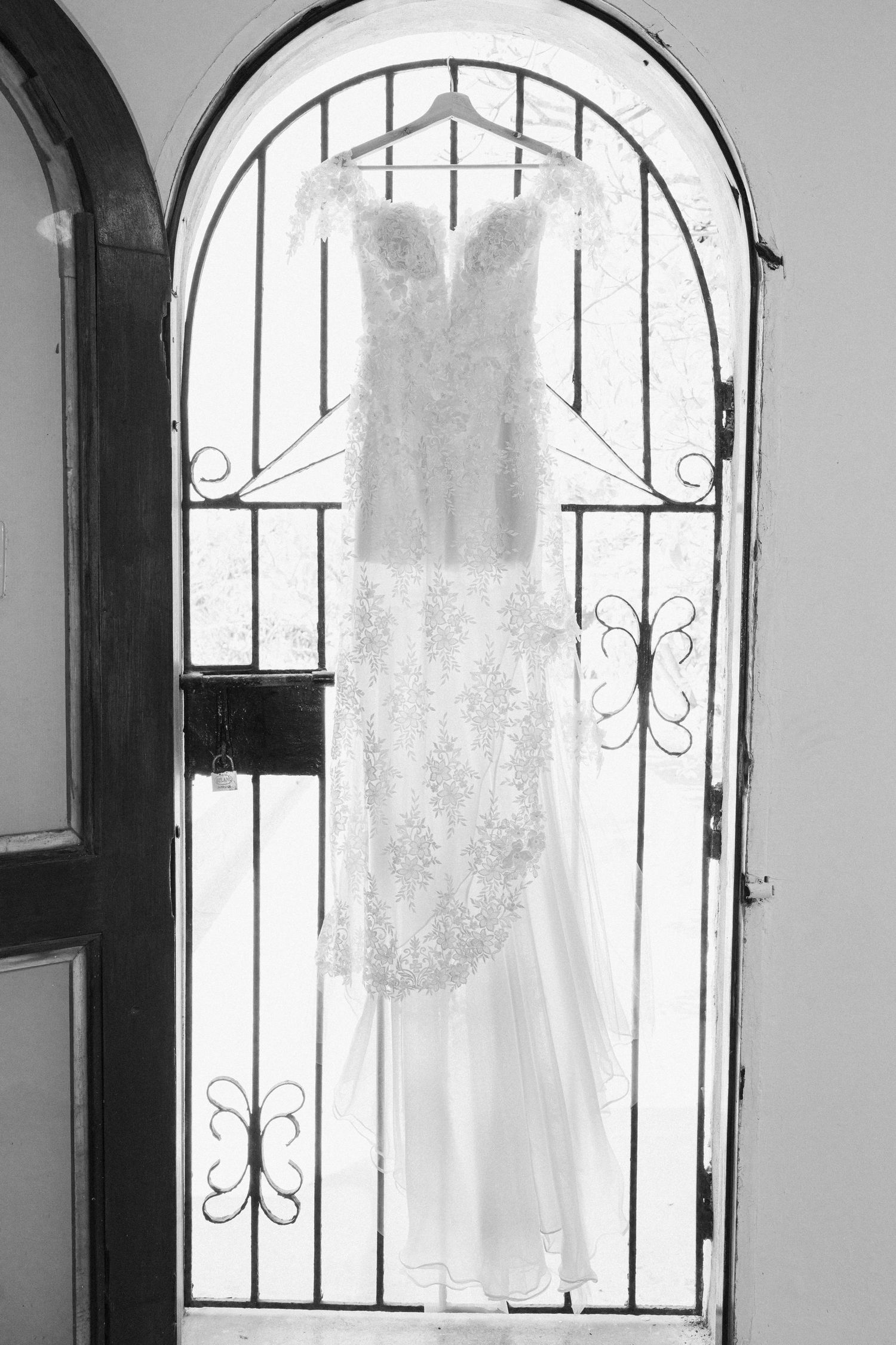 Black and white image of intricate lace wedding gown hanging on decorative wrought iron arched door.