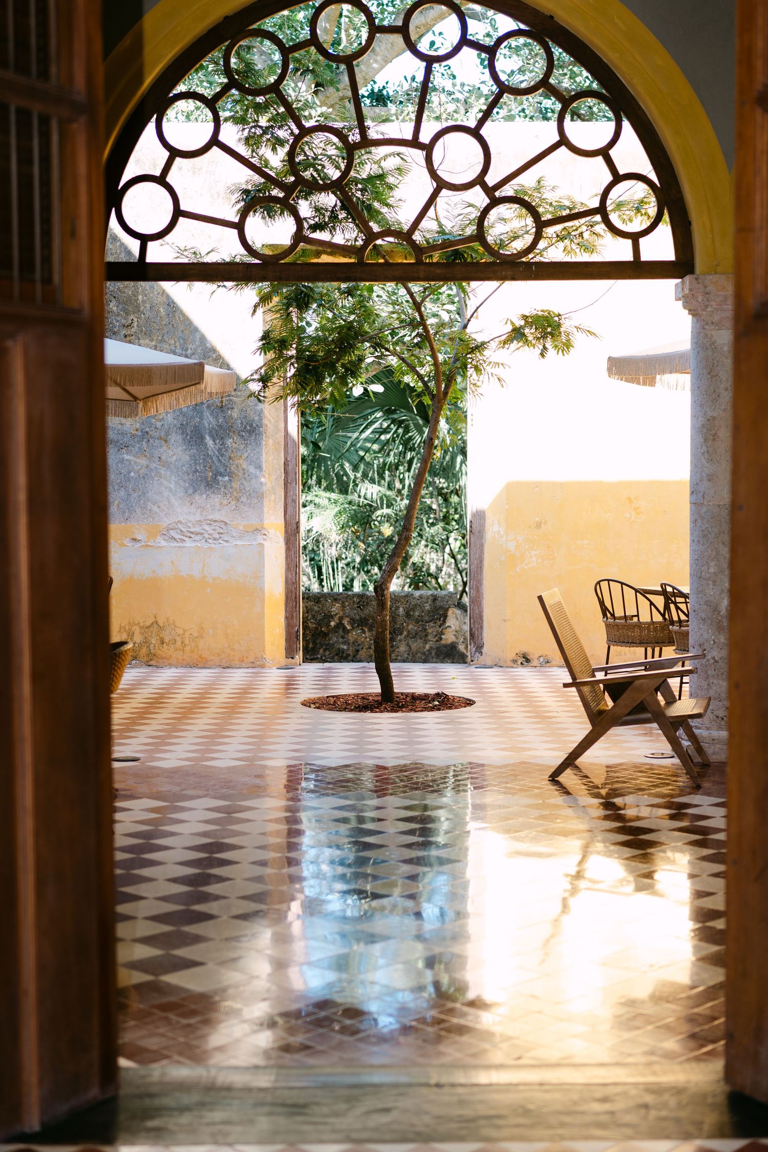 Sunlit courtyard viewed through ornate arched doorway with checkered floor and lone tree.