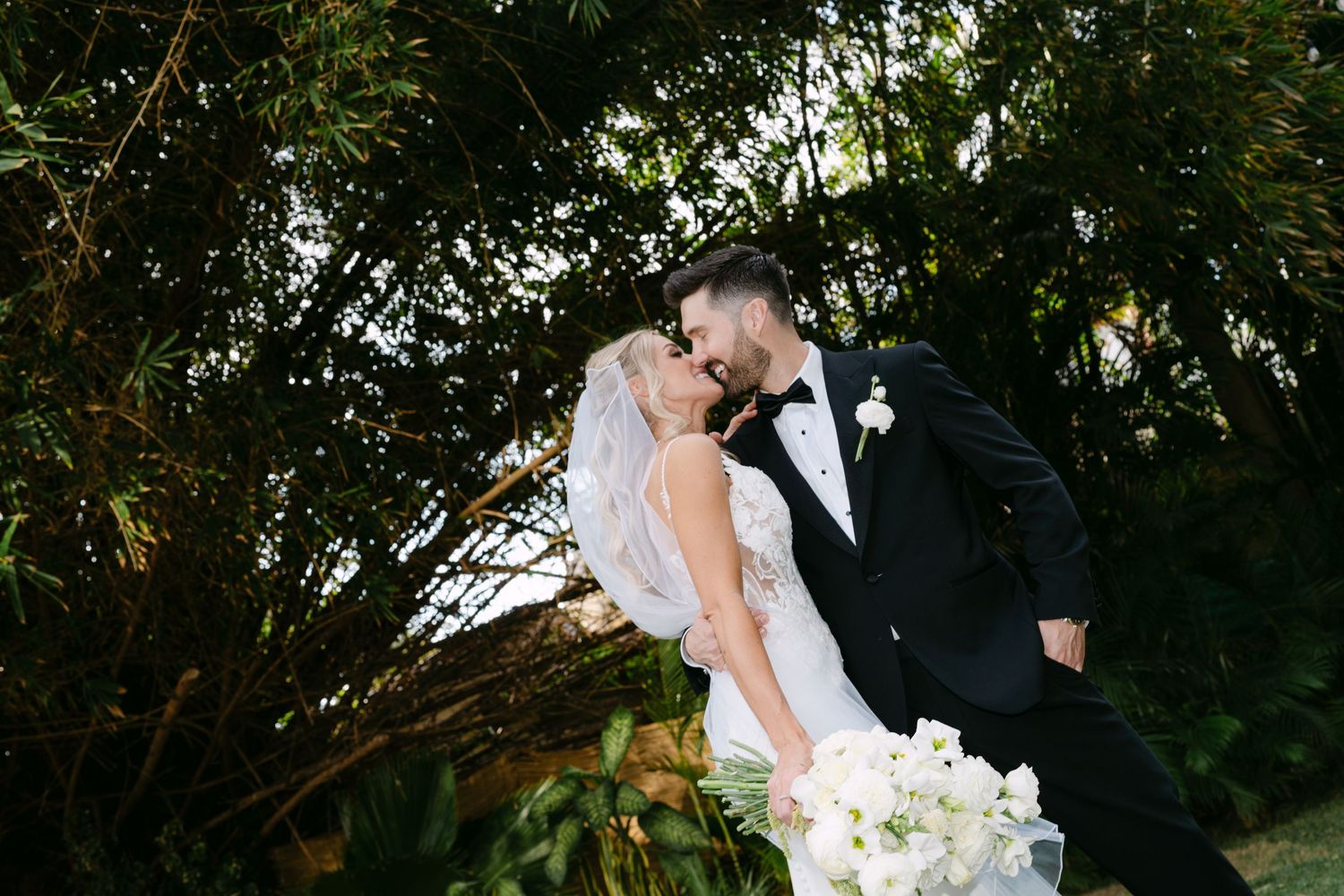 Bride and groom kissing surrounded by lush green foliage, bride holding white bouquet in elegant wedding attire.