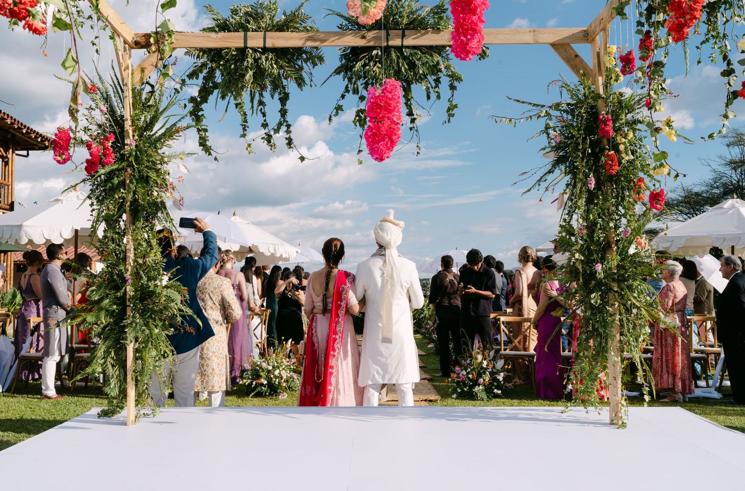 Indian wedding ceremony couple walks down white aisle under floral arch with colorful guests watching.
