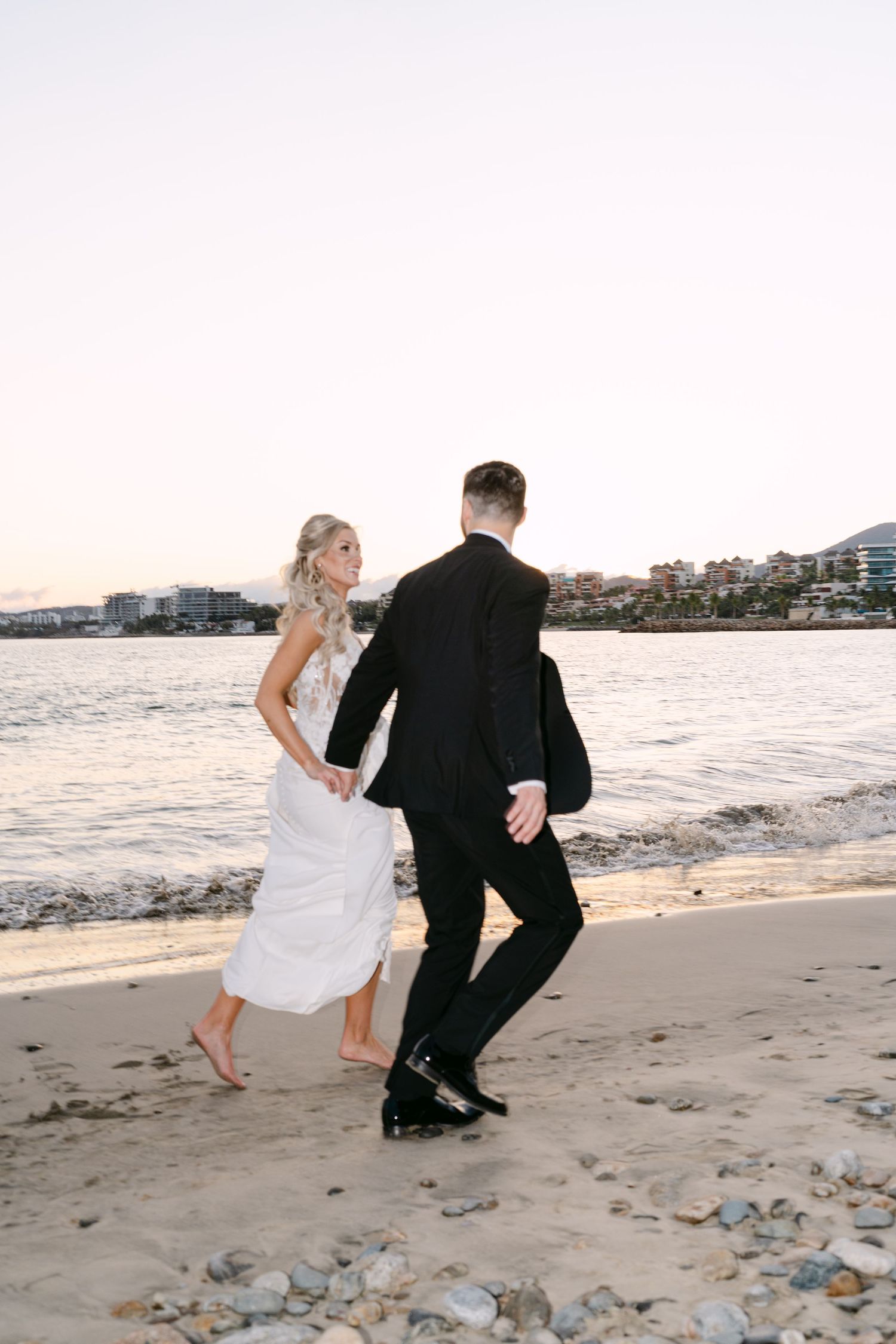Bride and groom walking hand in hand along a sandy beach at sunset.