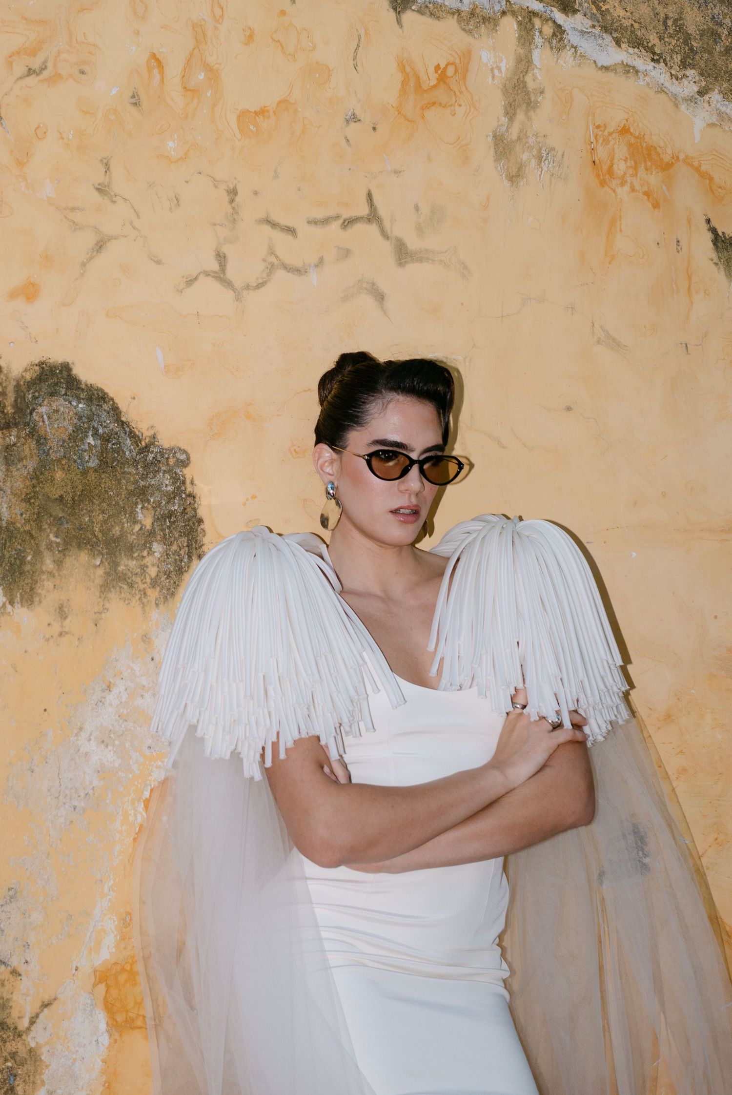 Bride in white fringe shoulder dress wearing sunglasses poses against rustic yellow wall.