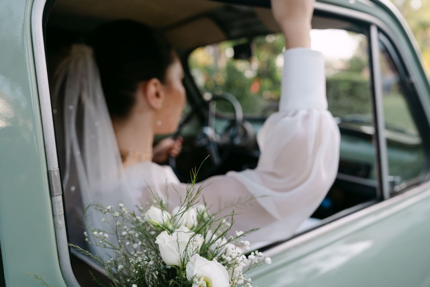 Bride in white dress holding bouquet of white flowers while sitting in a vintage mint green car on wedding day.