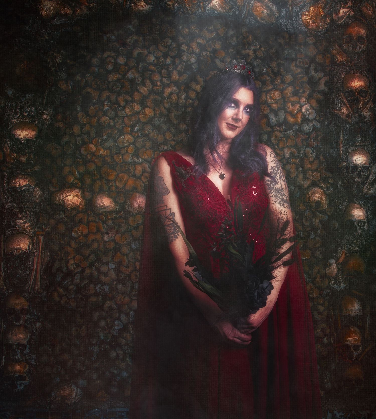 Woman with dark hair and arm tattoos in a flowing deep red gown holds dark feathers before a wall of skulls and bones.