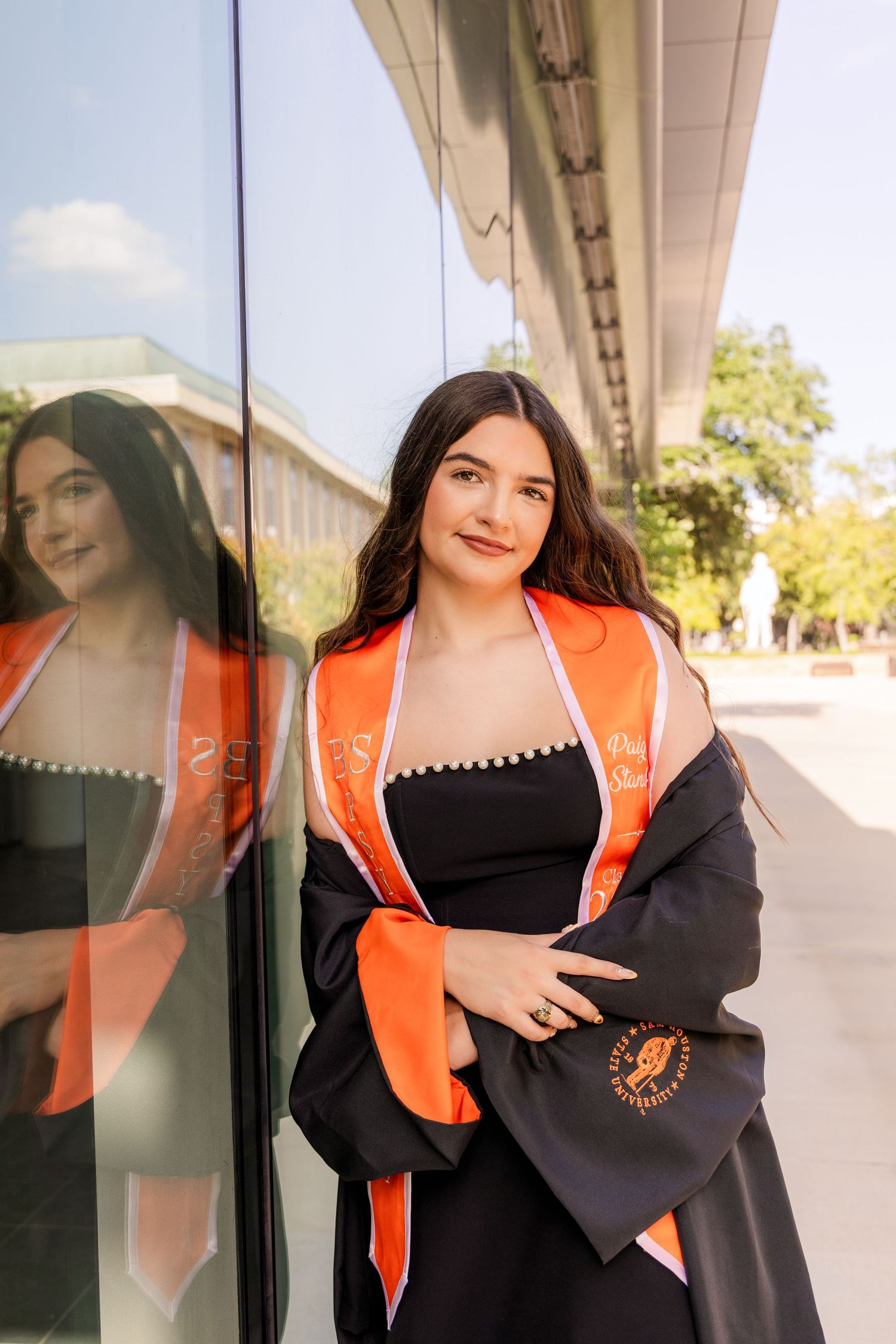 Graduate in a black dress, black gown and orange stole leans against a glass wall with her reflection beside her.