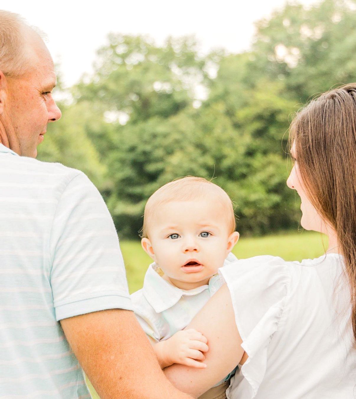Smiling baby in a white collared outfit looks at the camera while held by two adults outdoors in front of green trees.
