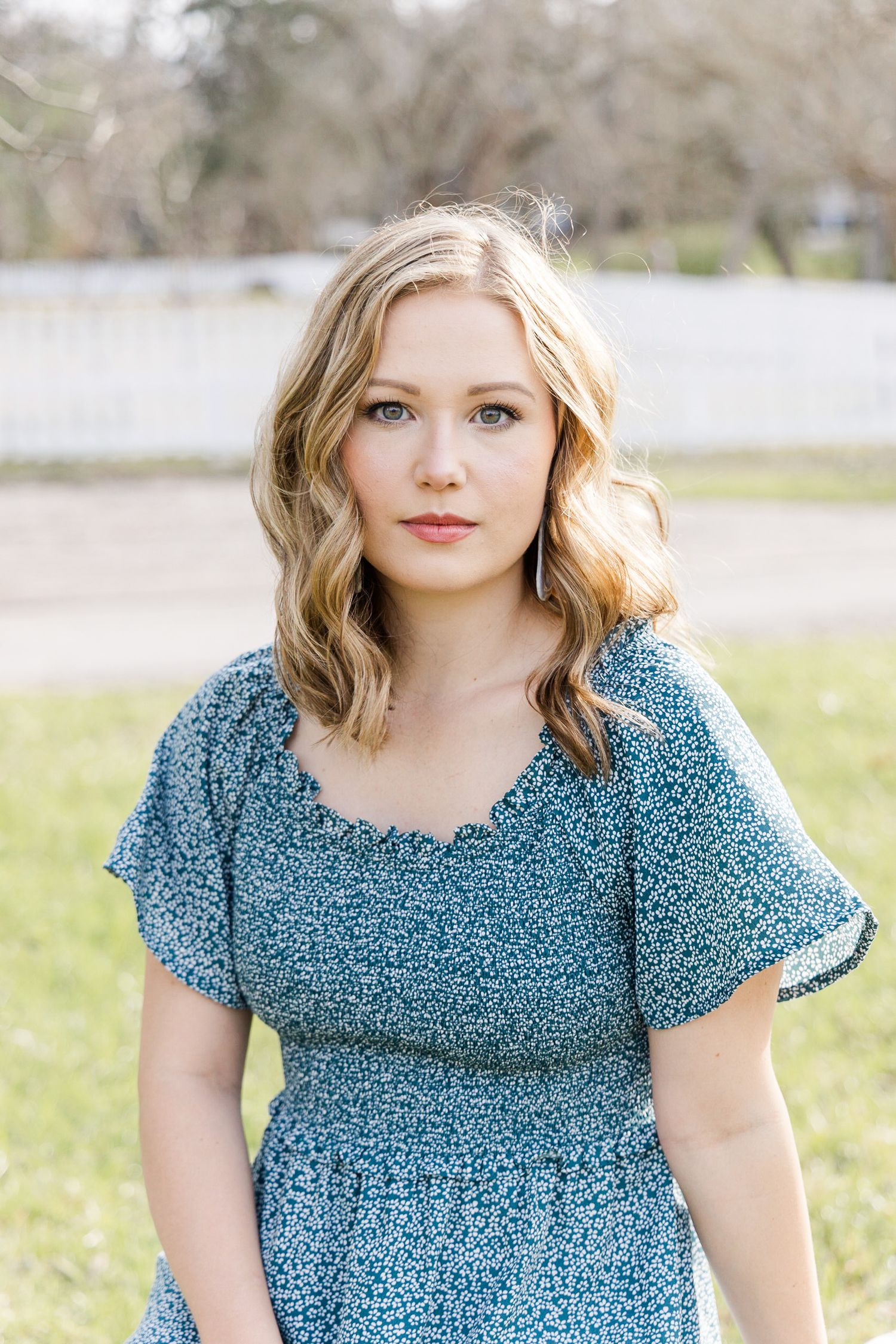 Young woman with wavy blonde hair in a blue floral ruffled dress poses outdoors in front of a white fence and grassy field.