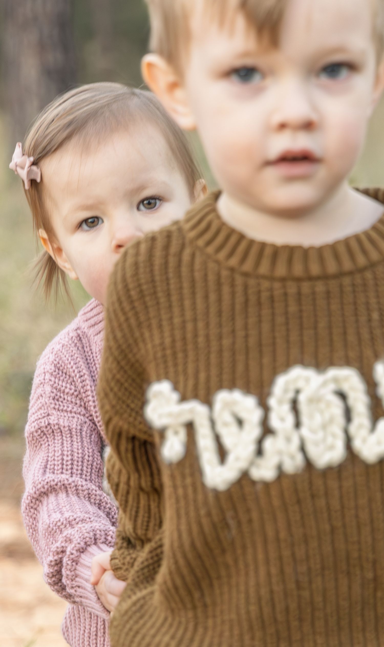 Two toddlers outdoors, one in a pink knit sweater with a bow peeking behind another in a brown embroidered sweater.