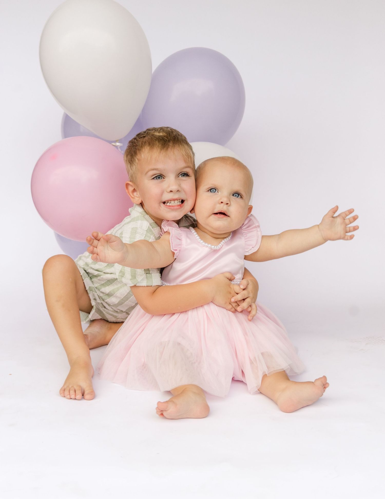 Two smiling siblings, a toddler boy hugging a baby girl in a pink tutu dress, sit in front of pastel pink, lavender and white balloons.