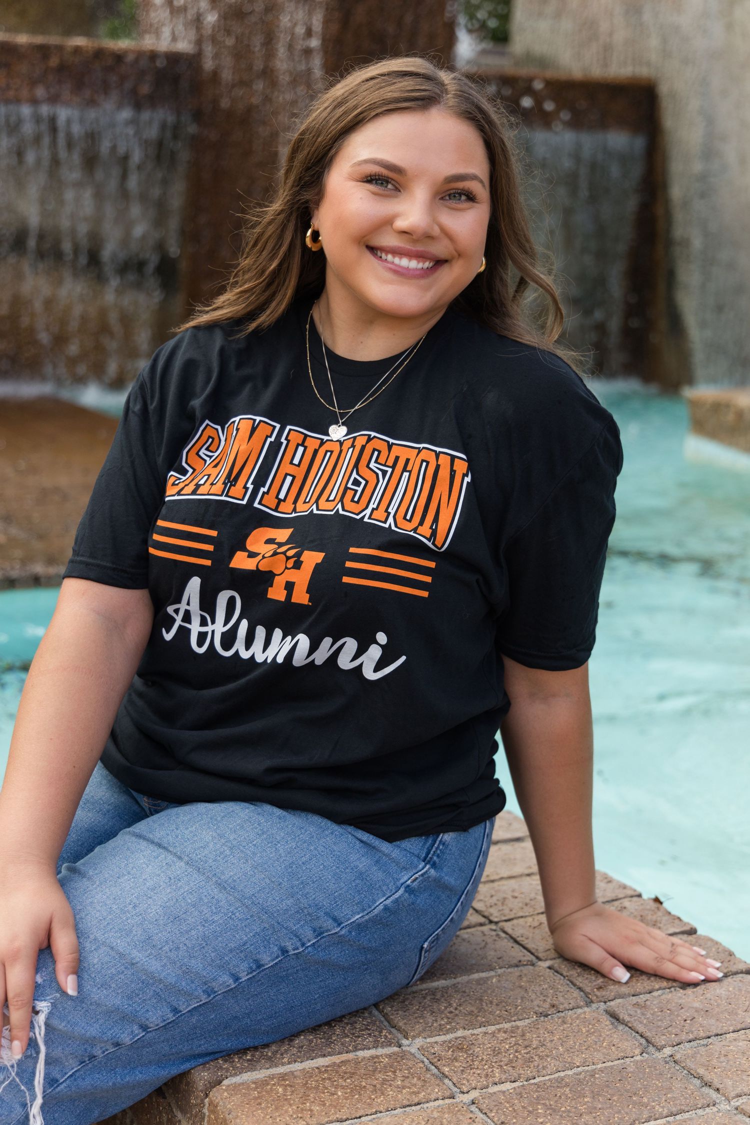 A smiling woman in a black Sam Houston Alumni t-shirt and jeans sits on a ledge beside a water fountain.