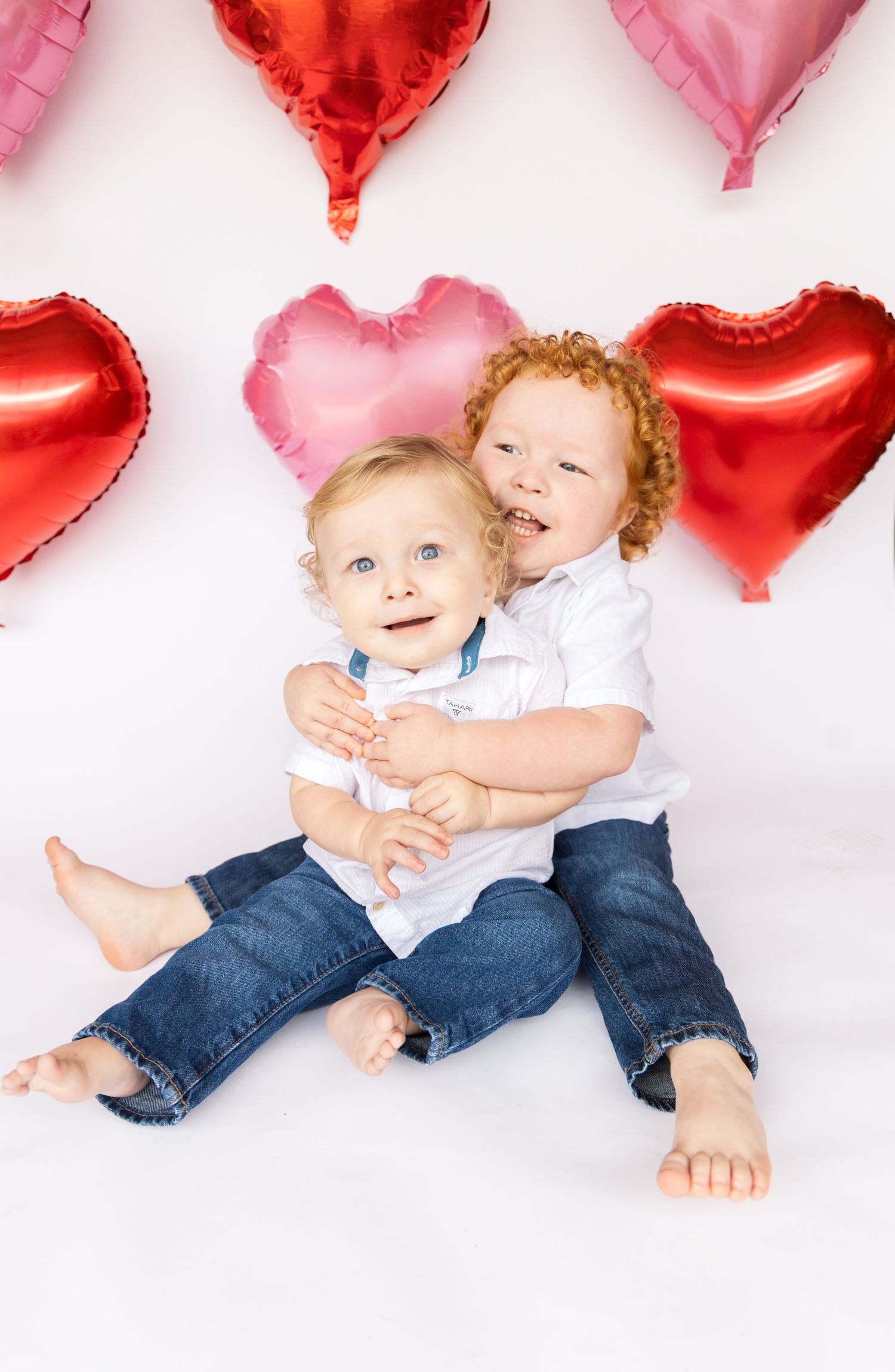Two smiling toddlers in white shirts and jeans sit together hugging in front of red and pink heart-shaped balloons.