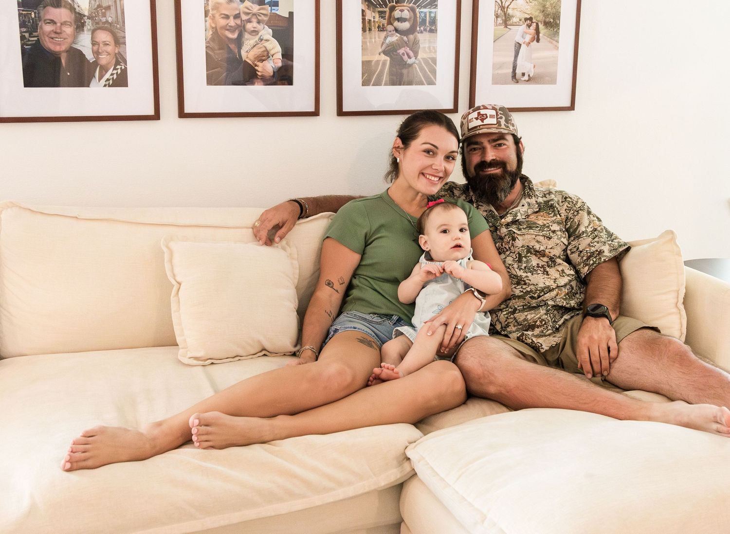 A smiling couple relaxes on a cream sofa with their baby beneath a wall of framed family photos.