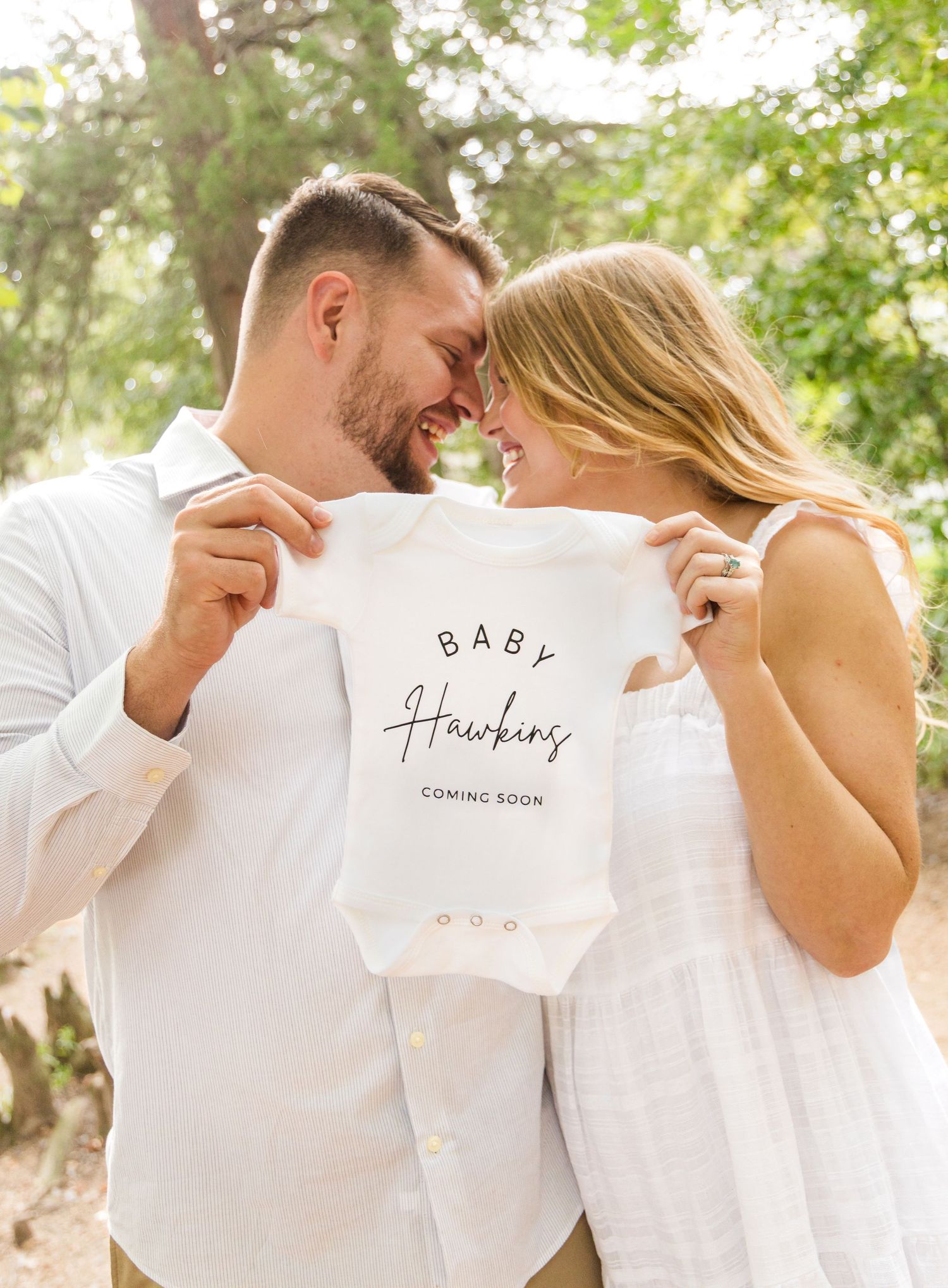 Smiling couple in white outfits embrace outdoors while holding a Baby Hawkins coming soon onesie to announce their pregnancy.