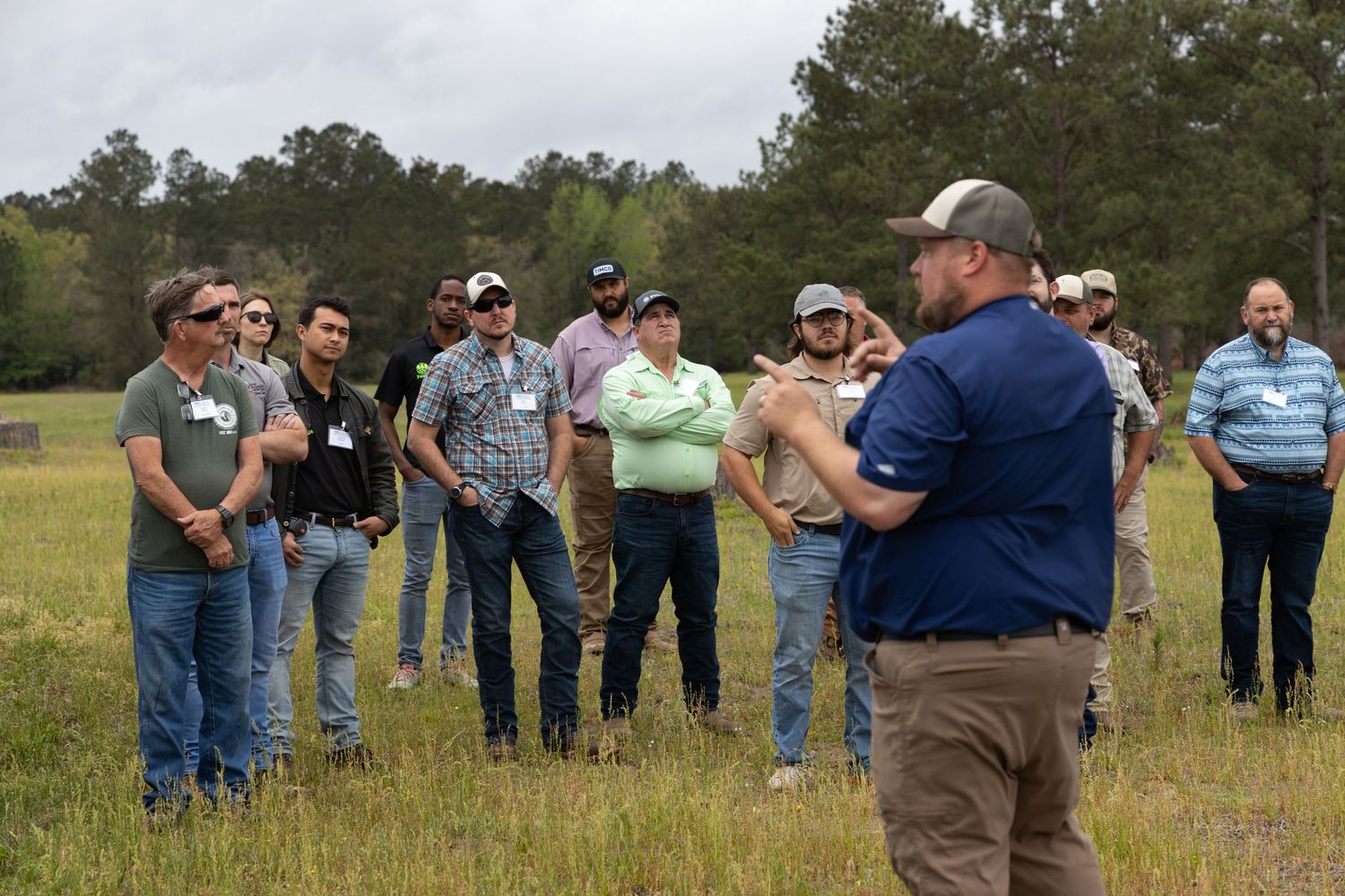 Man in a blue shirt and cap speaks to a group of attendees standing in an open grassy field with pine trees behind them.