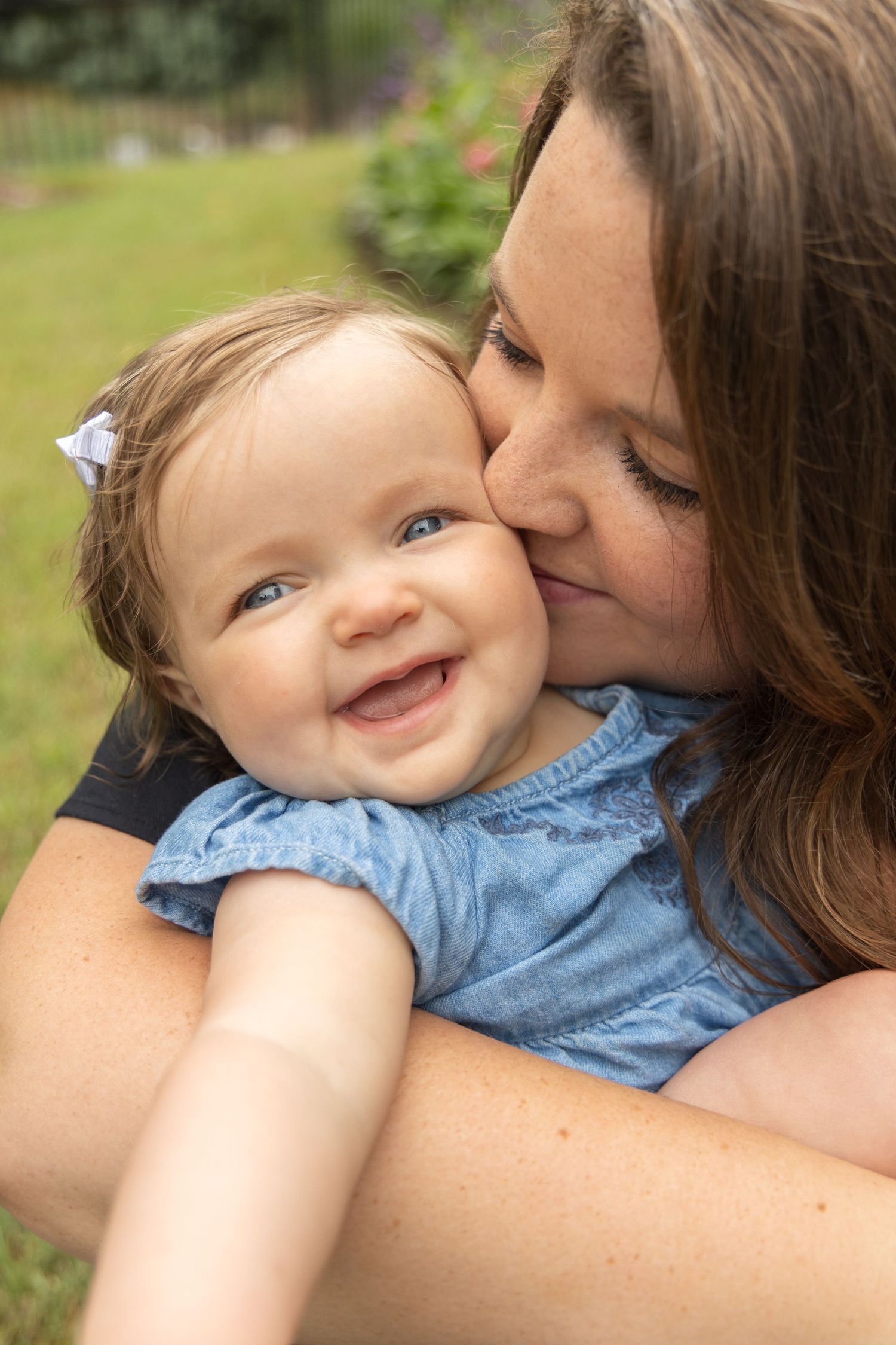 Mother kisses the cheek of her smiling baby girl in a blue denim outfit with a bow, in a green garden.